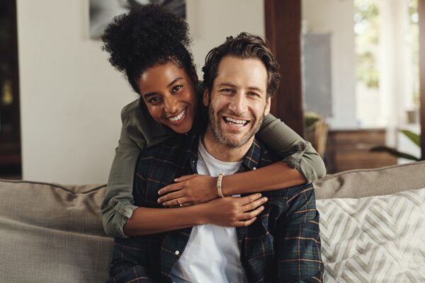 Happy, hug and couple with portrait on sofa for weekend bonding, interracial and romance together. Man, smile and woman with affection at home on couch for connection, love and support in marriage