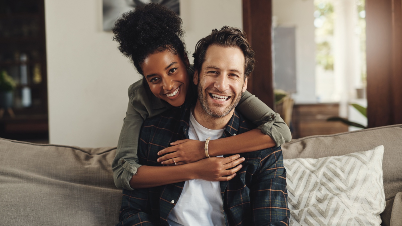 Happy, hug and couple with portrait on sofa for weekend bonding, interracial and romance together. Man, smile and woman with affection at home on couch for connection, love and support in marriage