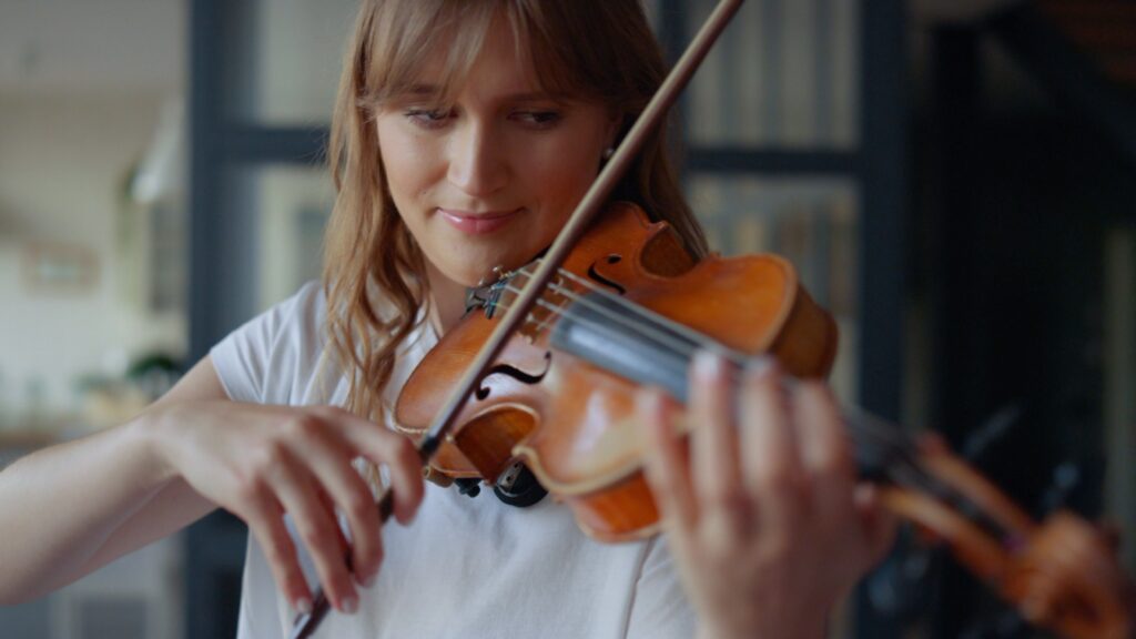 Young woman learning to play violin at home. Romantic girl playing violin with bow. Portrait of female musician performing on string instrument. Dreamy violinist fingers pressing strings on violin