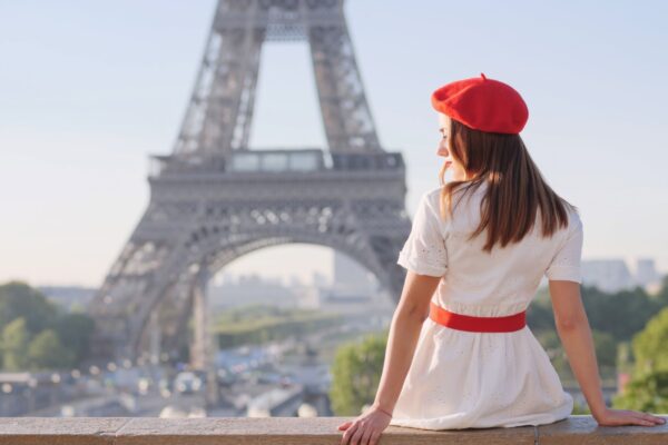 A beautiful girl in a white dress, a red beret sits against the backdrop of the Eiffel Tower in Paris. Travel to Paris. Ideas for a photo shoot.