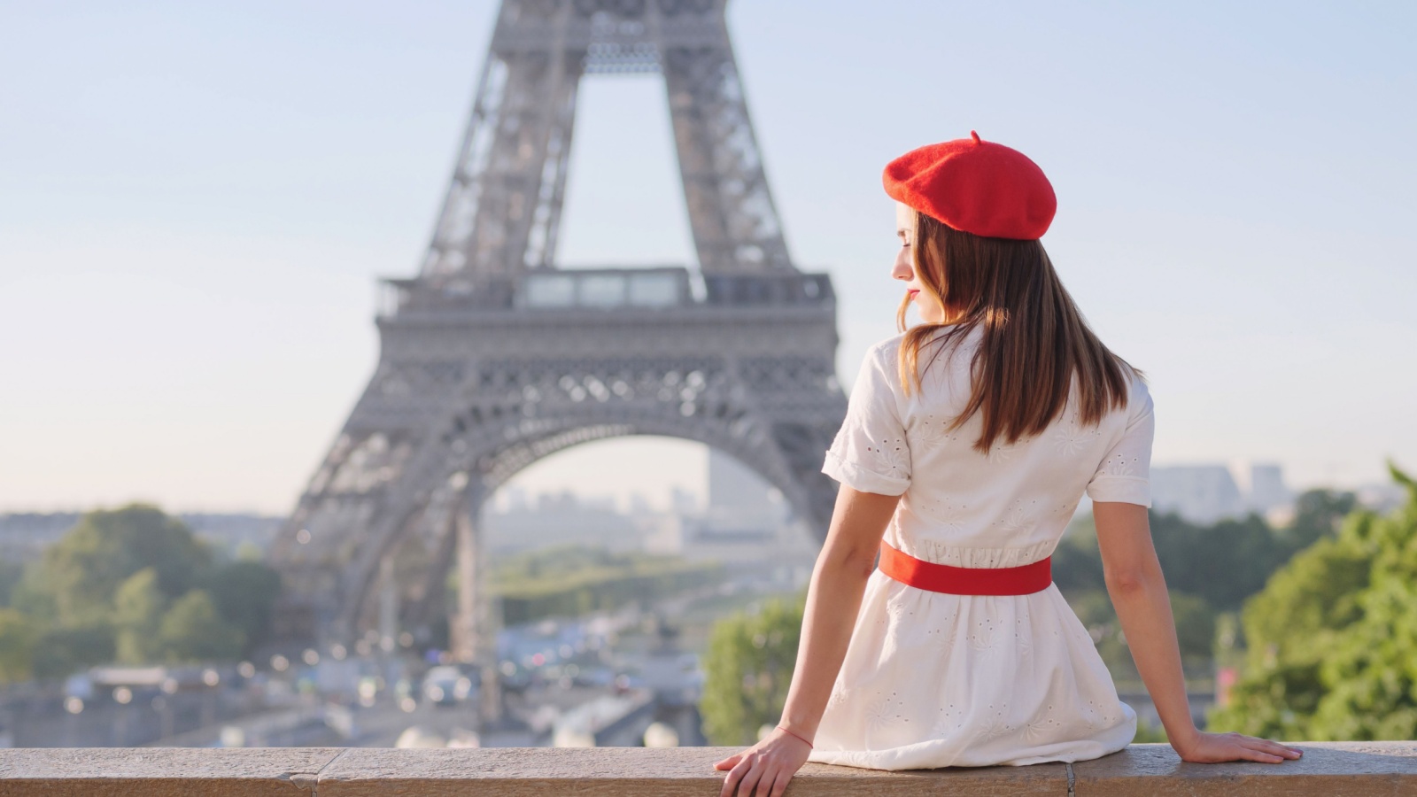 A beautiful girl in a white dress, a red beret sits against the backdrop of the Eiffel Tower in Paris. Travel to Paris. Ideas for a photo shoot.