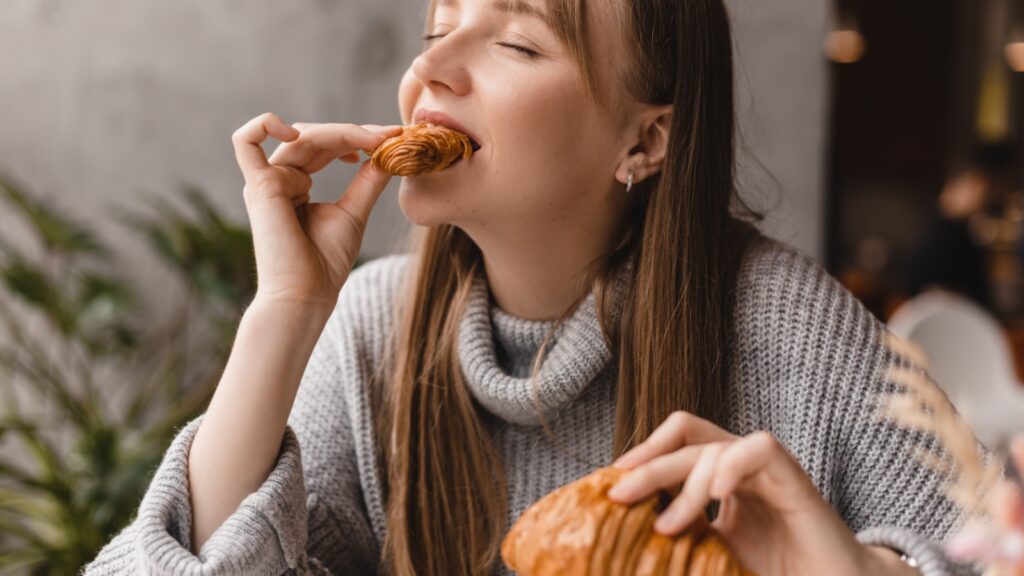 Young blonde woman with bang eating croissants at a cafe. Girl bite piece of croissant look joyful at restaurant. Cheat meal day concept. Woman is preparing with appetite to eat croissant.