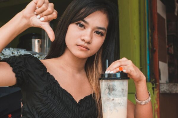 A dejected young asian woman gives the thumb down sign while sipping on milk tea. Possible negative review or rejection.