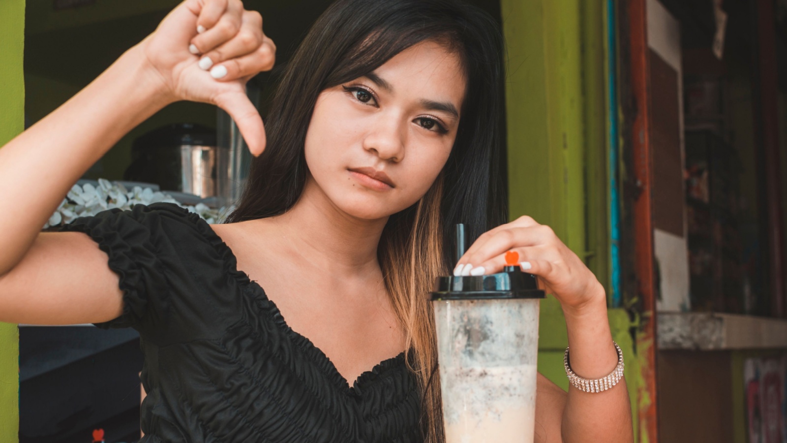 A dejected young asian woman gives the thumb down sign while sipping on milk tea. Possible negative review or rejection.