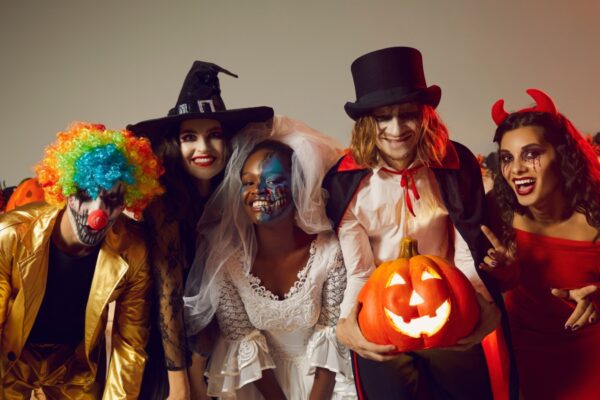 Happy young people dressed up as different spooky characters, with scary makeup on faces having fun at Halloween costume party. Group portrait of adult friends with traditional smiley jack-o-lantern