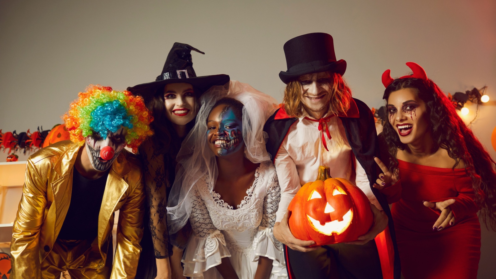 Happy young people dressed up as different spooky characters, with scary makeup on faces having fun at Halloween costume party. Group portrait of adult friends with traditional smiley jack-o-lantern