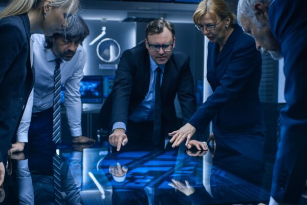 Diverse Team of Government Intelligence Agents Standing Around Digital Touch Screen Table and Tracking Suspect. FBI Agents Using Satellite Surveillance in the Dark Monitoring Room.