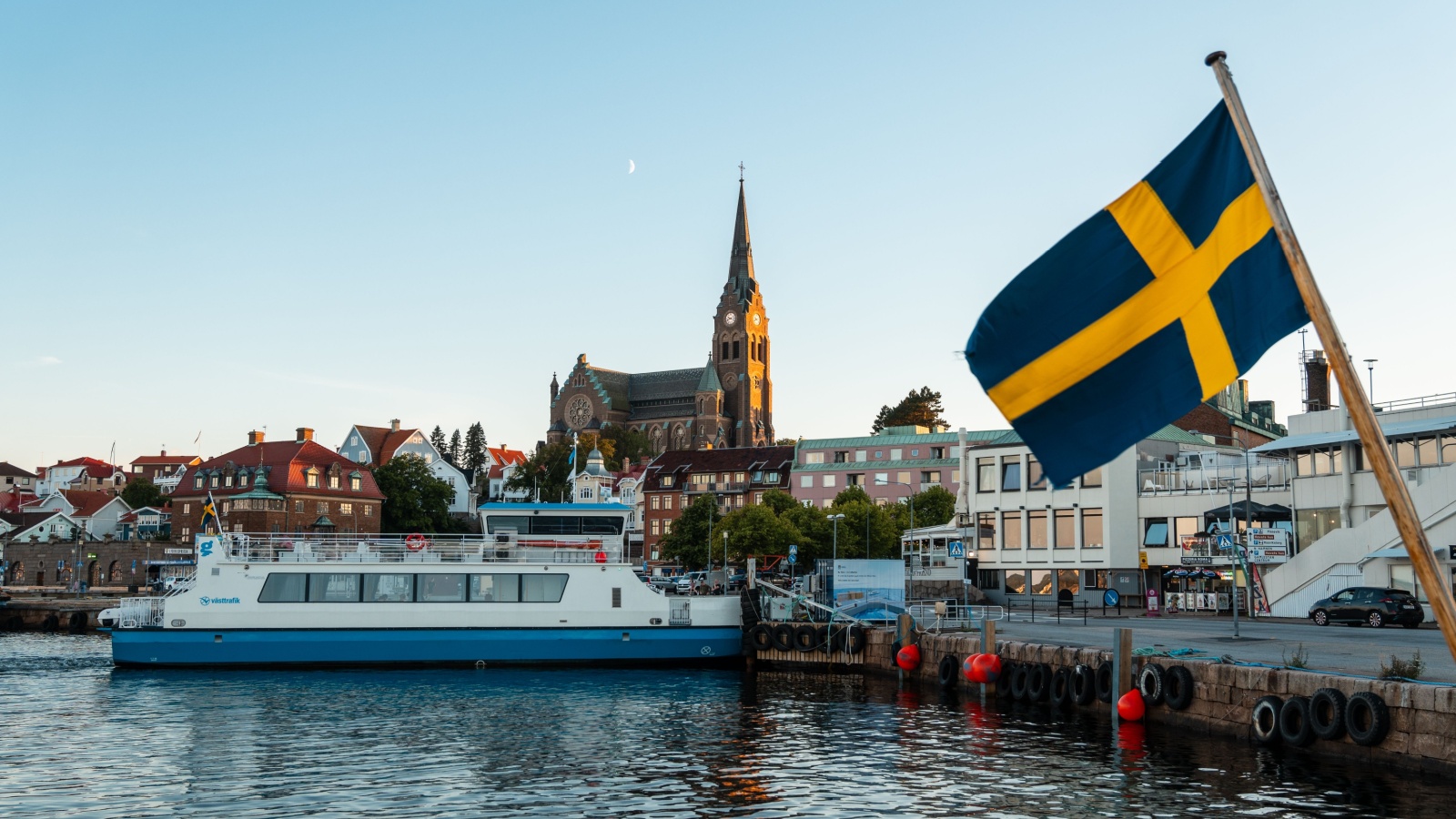 Lysekil, Sweden - July-12-2024, Ferry landing in the port with the church in the backgroundunder the moon in a midsummer night. A Sweden Flag waving in the foreground.