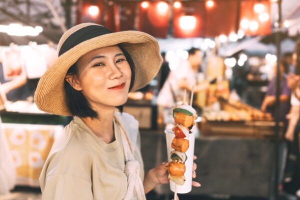 Poeple travel and eating street food concept. Happy young adult asian foodie woman holding bbq grilled skewers at outdoor vendor night market.