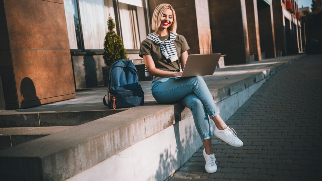 Full body photo of young blonde woman sit concrete border netbook wear khaki clothes tied shoulders sweater walk outside urban city park