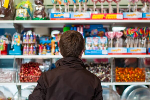 A young person with brown hair wearing a brown casual jacket seen from behind is standing in front of colourful shelves filled with sweets and candy in a store.